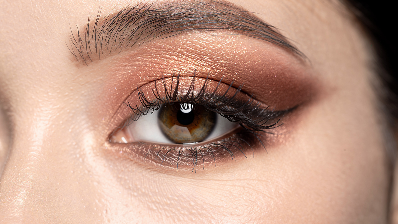 Gros plan d’un œil de femme maquillé avec un fard chrome cuivre rosé et un smokey eye brun diffus, sur une peau naturelle avec les cheveux attachés.