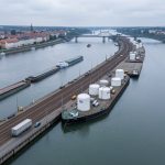 Vue aérienne réaliste du port fluvial de Strasbourg sur le Rhin, avec des barges, des voies ferrées et des citernes cylindriques d’hydrogène dans une ambiance industrielle sobre.