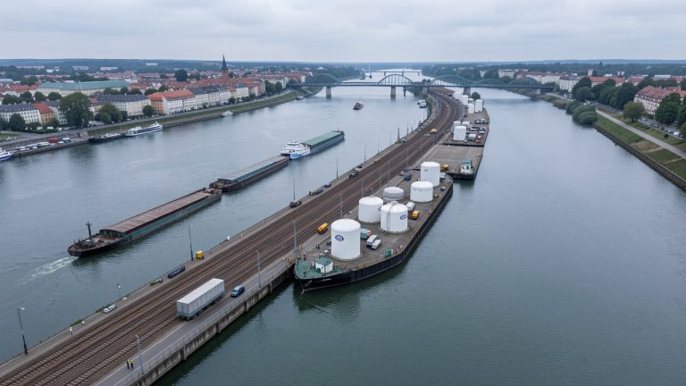 Vue aérienne réaliste du port fluvial de Strasbourg sur le Rhin, avec des barges, des voies ferrées et des citernes cylindriques d’hydrogène dans une ambiance industrielle sobre.